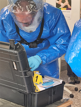 Photo of a simulation exercise: A man in a protective suit bends over a suitcase. (© RKI) Photo of a simulation exercise: A man in a protective suit bends over a suitcase.